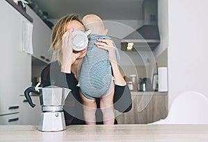 Modern tired mother and little child after sleepless night. Exhausted woman with baby is sitting with coffee in kitchen. Life of