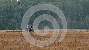 A modern tractor plows brown fertile soil on farmland next to a forest