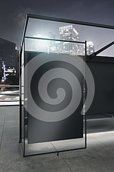 Modern vertical glass bus stop with empty black billboard and reflections on night city background. Mock up