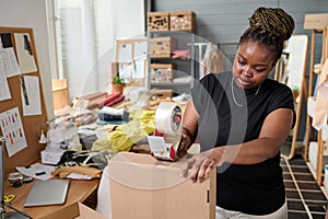 Modern warehouse worker sealing cardboard box with cellotape by workplace