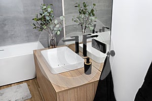 A modern washbasin in the bathroom in a wall-mounted cabinet with a matte black faucet, a visible mirror and artificial flowers.