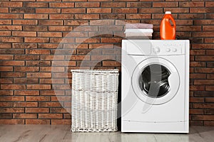 Modern washing machine with stack of towels, detergent and laundry basket near wall