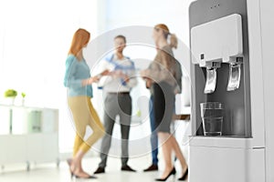 Modern water cooler with glass and blurred office employees on background