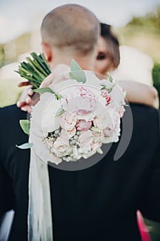 Modern wedding bouquet in bride hands. Gorgeous bride gently hugging groom in sunset light. Beautiful stylish wedding couple