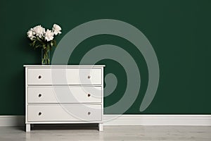 Modern white chest of drawers with beautiful bouquet near green wall indoors. Space for text