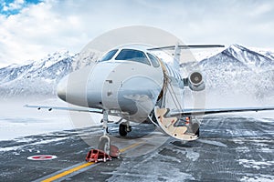 Modern white executive aircraft with an opened gangway door at the winter airport apron on the background of high scenic snow