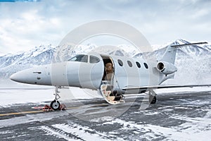 Modern white executive jet plane with an opened gangway door at the winter airport apron on the background of high scenic snow