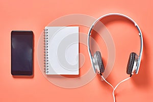 Modern white headphones with cable near smartphone or tablet and blank paper notepad on orange background