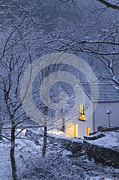 Modern white house with light windows surronded by mountain, snow and trees