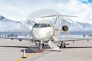 Modern white private jet with an opened gangway door at the winter airport apron on the background of high mountains