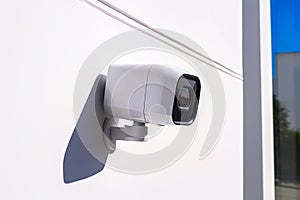 A modern white security camera mounted on an outdoor wall in bright sunlight.