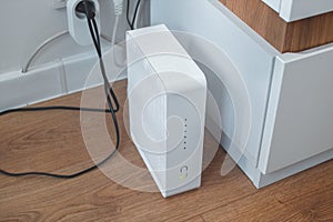A modern Wi-Fi router plugged into an outlet, white and without antennas, stands on the floor in a home room.
