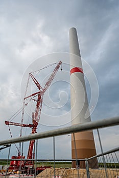 Modern wind turbine construction site with crane next to it and metal fence in front during cloudy day, view from low angle