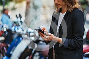 Modern woman in business suit holding in hands smartphone uses app scooter rental service outside in city, female manager looks