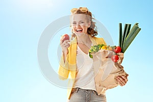 Modern woman with paper bag with groceries biting apple ag
