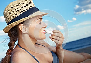 Modern woman on seashore applying sun protection lipstick