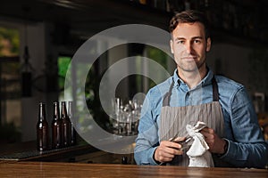 Modern work of bartender. Handsome man in apron with stylish haircut, rubs glass