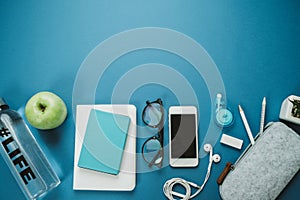 Modern Work space for healthy person: pencils, water, apple, phone, glasses, headphones on a blue background. Top view, flat lay
