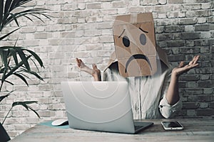 Modern worker with carton sad box on head open arms with desolation gesture in front of a laptop. Online crypto smart working