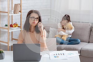 Modern working mom doing online job, her neglected daughter hugging teddy bear, feeling lonely and sad at home