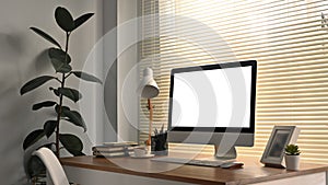 Modern workplace interior, white blank screen computer monitor, picture frame, lamp and books on wooden desk