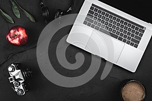 Modern workplace with a laptop keyboard, cup of coffee, red apple, photo camera and headphones on a dark office desk background