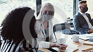 Modern workplace meeting after COVID-19 lockdowns. Young female multiethnic partners talk at office wearing face masks.