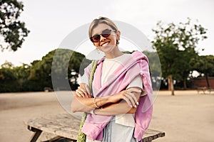 Modern young caucasian girl smiles teeth looking at camera standing outdoors with arms crossed.