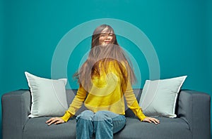 Modern young girl shaking her long brown tousled hair, enjoying haircare result, sitting on sofa. Natural hairs beauty