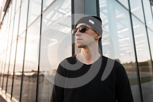 Modern young handsome hipster man in stylish knitted black hat in trendy trendy sunglasses in a t-shirt posing