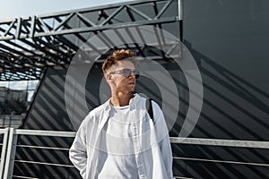 Modern young hipster man in stylish white clothes in trendy sunglasses in a black vintage bag is standing near a modern building