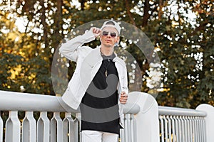 Modern young hipster man in a summer white jacket in sunglasses in a trendy jeans t-shirt poses in a park near a metal fence.