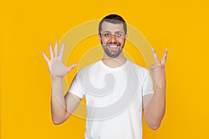 Modern young man with a beard in a white tank top shows number seven with fingers on hand, smiling confidently and happily,