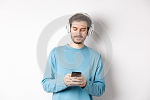 Modern young man in blue sweatshirt put on music on smartphone, listening songs in wireless headphones, white background