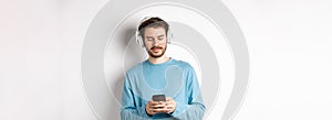 Modern young man in blue sweatshirt put on music on smartphone, listening songs in wireless headphones, white background