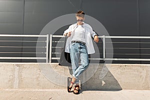 Modern young man hipster in fashionable summer white clothes in sunglasses in stylish red leather sandals with a bag stands