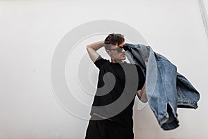 Modern young man hipster puts on a stylish denim jacket. Attractive guy in fashionable clothes in sunglasses posing on a street in