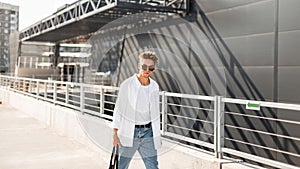 Modern young man hipster with stylish hairstyle in fashionable summer clothes with a black cloth bag in vintage sunglasses walks