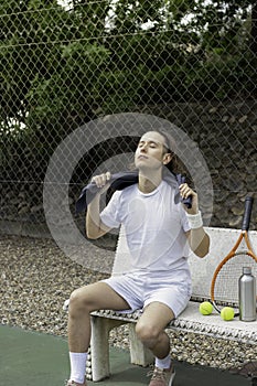 Modern young man with long hair sitting exhausted with a towel around his neck after playing a game of tennis