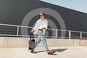 Modern young man in stylish clothes in sunglasses in red leather sandals with a vintage black bag travels on a bright sunny day