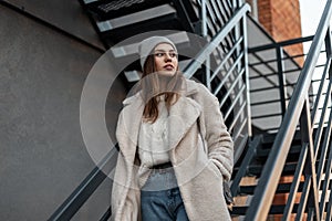 Modern young model of a stylish woman in a fashionable faux fur coat in a trendy knitted hat in a sweater in jeans stands near a