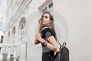 Modern young woman in black fashionable clothes straightens hair. Attractive joyful girl model in a black dress with a backpack