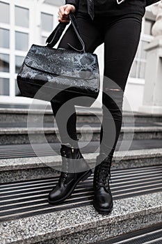 Modern young woman with a fashionable black leather bag in jeans in autumn stylish leather black boots is standing on the street.