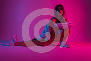 Modern young woman in fashionable clothes is doing stretching indoors with neon bright pink light. Attractive girl posing in a