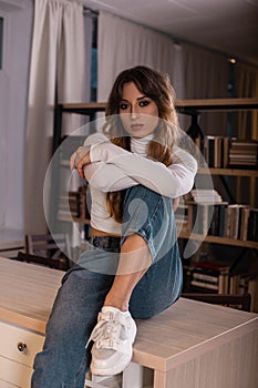 Modern young woman model in a fashionable white t-shirt in vintage blue jeans in youth sneakers sits on the kitchen table indoors