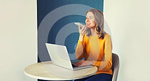 Modern young woman working with laptop talking on smartphone using voice command recorder, assistant or takes calling on her desk