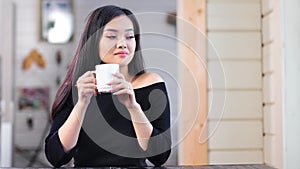 Modest adorable Asian young girl relaxing sitting at table and drinking hot beverage at home