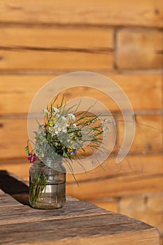 Modest bouquet of white and pink wildflowers with green twigs in glass jar is placed on wooden table in oudoor. photo