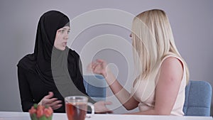 Modest Muslim woman in hijab talking with blond Caucasian friend criticizing her outfit. Conservative lady and modern