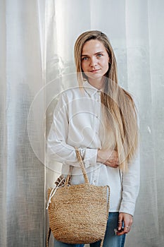 Modest woman in white sunlit room posing for a photo, holding wicker bag